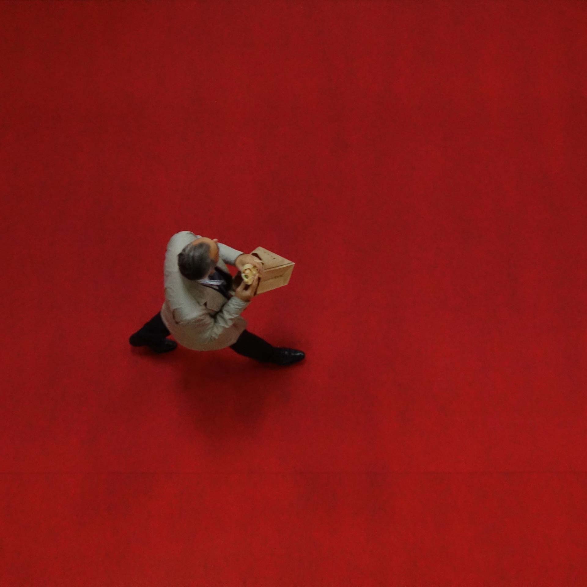 Vue en plongée d’une personne marchant sur un sol rouge uni en tenant dans les mains un petit sachet en papier, illustrant un mouvement dynamique dans un environnement structuré.
